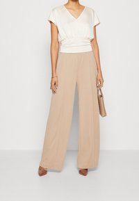 Krémová blůza s výstřihem do V a nařaseným pasem, spojená s béžovými širokými kalhotami s vysokým pasem. Hnědé boty na podpatku a tkaná taška.