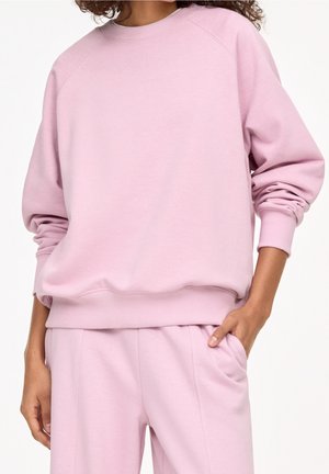 Personne portant un sweat rose pastel et un pantalon assorti, une main dans la poche, debout devant un fond blanc uni.
