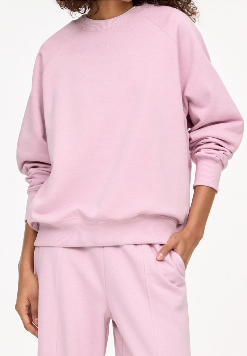 Personne portant un sweat rose pastel et un pantalon assorti, une main dans la poche, debout devant un fond blanc uni.