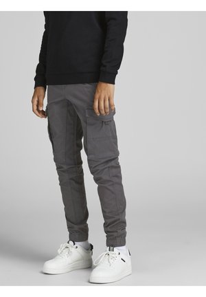 Mand iført grå cargobukser med flere lommer, hvide sneakers og en sort langærmet top stående på en lysegrå baggrund.