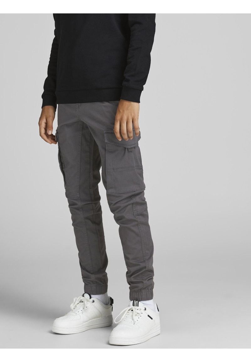 Homme portant un pantalon cargo gris à plusieurs poches, des baskets blanches et un haut noir à manches longues, debout sur un fond gris clair.