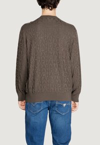 Maglione marrone a maglia con motivo a logo in rilievo. Maniche lunghe, polsini e orlo a costine, indossato con jeans blu. Design casual.