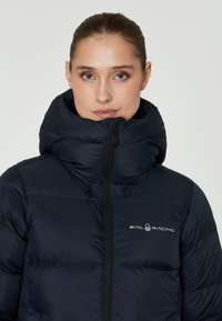 Marineblaue Pufferjacke mit hohem Kragen, Reißverschluss vorne und Logo auf der Brust. Sie verfügt über ein gestepptes Design und strapazierfähigen Stoff.