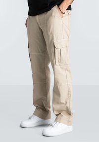Beige cargo pants feature multiple pockets, a relaxed fit, and stitched detailing. Paired with white sneakers, the fabric appears durable.