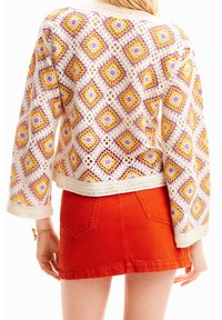 Cardigan en crochet avec un motif losange coloré en nuances de violet, jaune et crème, assorti à une jupe en jean rouge.