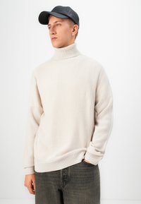 Young man with light beige turtleneck sweater, black cap, and dark jeans standing against a plain white background, looking left.