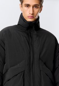 Schwarze isolierte Jacke mit hohem Kragen, ausgestattet mit einem durchgehenden Reißverschluss und schrägen Fronttaschen. Der Stoff hat eine weiche, strukturierte Oberfläche.