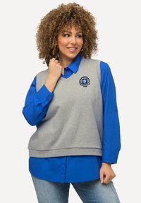 Lys grå ærmeløs sweater vest med v-hals, lagd over en lys blå langærmet skjorte. Har et broderet logo på vesten.