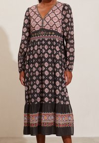 Kvinna klädd i en svart midiklänning med rosa och orange geometriska mönster, V-ringning, långa ärmar samt broderad midja och fåll.