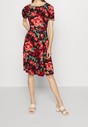 Robe florale à manches courtes, base noire, motif de grandes fleurs rouges et vertes, ceinture à la taille, longueur au genou, associée à des sandales à talons blocs de couleur beige clair.