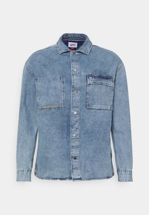 Chemise en denim bleu clair à manches longues, avec boutons sur le devant, deux poches poitrine et marquage Tommy Jeans sur la poche gauche et l’étiquette du col.
