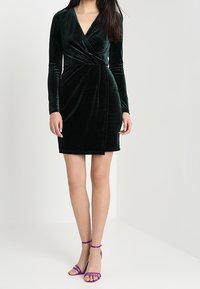 Kvinde iført en sort velour langærmet omslagskjole og lilla højhælede sandaler, som står mod en ensfarvet lys baggrund.