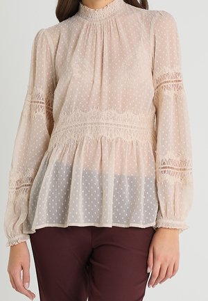 Blusa beige a pois con maniche lunghe, colletto alto, pizzo in vita e orlo svasato. Realizzata in tessuto leggero e trasparente.