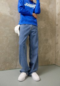 Blå sweatshirt med "JORDAN"-logotyp, kombinerad med lösa blå denimjeans och vita sneakers, mot en betongbakgrund.
