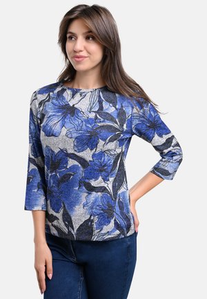Bluse mit blauem und grauem Blumenmuster, Dreiviertelärmeln, rundem Ausschnitt und weicher Textur. Kombiniert mit dunklen Jeans.