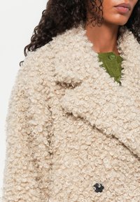 Beige fluffig kappa med en texturerad, lockig yta; med dubbelknäppt design och en svart knapp. Grön stickad lager synligt under.
