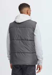 Grå puffervest laget av kviltet materiale, med høy krage og sett fra baksiden. Brukt over en grå hettegenser, med en løs passform.