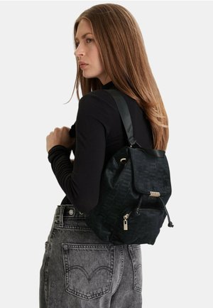 Donna con capelli lunghi castani, indossa una maglietta nera e jeans grigi, porta uno zaino nero Guess su una spalla.