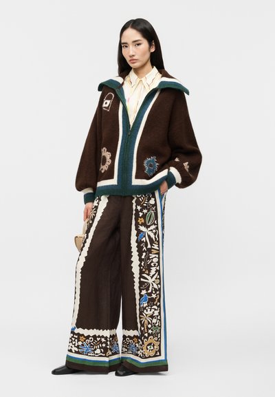 Γυναίκα που φορά καφέ φαρδιά τζάκετ με μπλε και κρεμ μοτίβα, λευκό και κίτρινο πουκάμισο, και παντελόνια με φαρδιά μπατζάκια και πολύχρωμες περιφερειακές διακοσμήσεις.