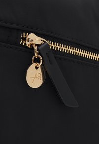 Detail einer schwarzen Nylon-Tasche mit einem goldenen Reißverschluss, einem runden goldenen Anhänger mit eingeprägtem Logo und einem schwarzen Ziehband.