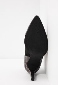 Chaussure à talon haut à bout pointu avec semelle noire texturée et dessus gris à motif peau de serpent, positionnée verticalement contre un coin blanc.