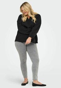Černý strukturovaný top s výstřihem do V a dlouhými rukávy, kombinovaný se šedými skinny džíny a černými balerínami. Neutrální pozadí.