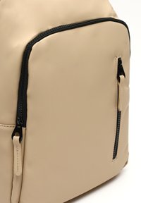 Béžový batoh vyrobený z hladkého syntetického materiálu, s čiernymi zipsovými akcentmi a štruktúrovaným dizajnom s viacerými zipsovými vreckami.