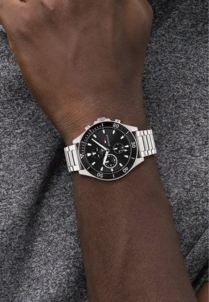 Zwarte chronograaf horloge met zilveren metalen band, ronde kast en luminescente markeringen, weergegeven op een pols tegen een grijze achtergrond.