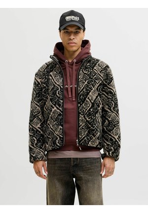 Mann trägt schwarze und beige gemusterte Fleecejacke mit Reißverschluss über einem bordeauxfarbenen Hoodie und schwarzer Kappe mit dem Schriftzug „Originals“, steht vor einfarbigem Hintergrund.