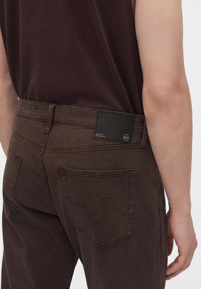Homme portant un jean marron foncé et un t-shirt noir, vu de dos au niveau de la taille avec un patch noir estampillé sur le jean.