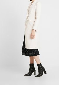 Abrigo largo de color beige claro con un diseño a medida, que cuenta con un solo botón y bolsillos laterales. Se lleva sobre un vestido negro con lunares y botas de tobillo negras.
