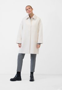 Weißer Wollmischungs-Mantel mit traditionellem Kragen, Knopfverschluss vorne, Seitentaschen und lockerer Passform. Kombiniert mit grauen, maßgeschneiderten Trousers und schwarzen Stiefeln.