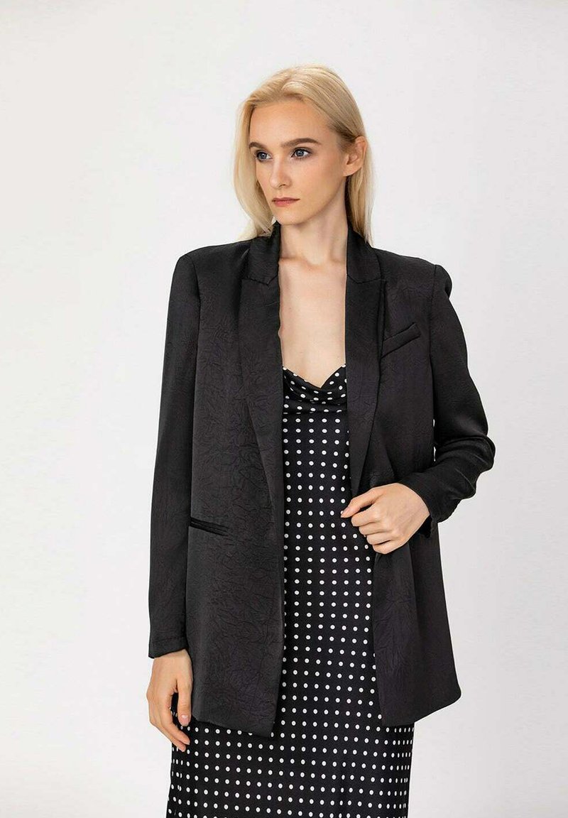 Blazer noir texturé avec revers, deux poches avant. Porté sur une robe à pois noir et blanc, mettant en valeur une silhouette superposée.