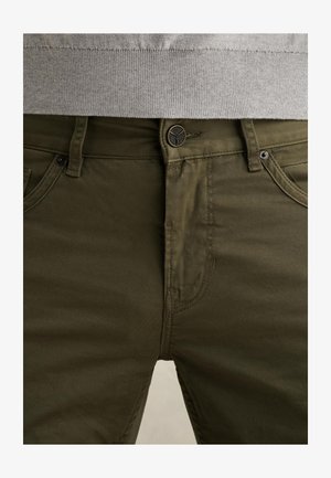 Detailný záber na olivovo zelené nohavice s gombíkom a zipsom na chrbte, nosené pod svetlo sivým svetrom.