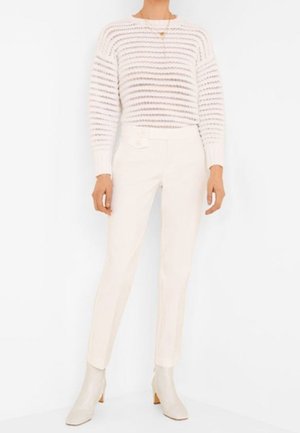 Žena ve světlém texturovaném pleteném svetru, s vysokým pasem bílých kalhot na míru a bílých kotníkových botách stojí před jednobarevným pozadím.