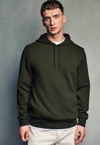 Grønne mønstrede hættetrøje med en tekstureret overflade, der har snørehætte og ribbede ærmer, lagdelt over en hvid T-shirt.