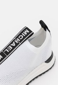 Weißer Sneaker mit atmungsaktivem Mesh-Obermaterial, schwarzem elastischem Riemen mit der Aufschrift "MICHAEL" am Knöchel und einer schwarzen und weißen flexiblen Sohle.