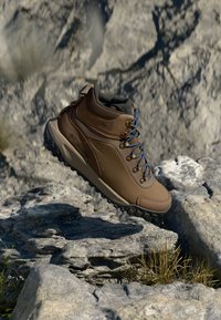 Brown hiking boot with a textured synthetic upper, padded collar, blue laces, and rugged outsole, set against rocky terrain.