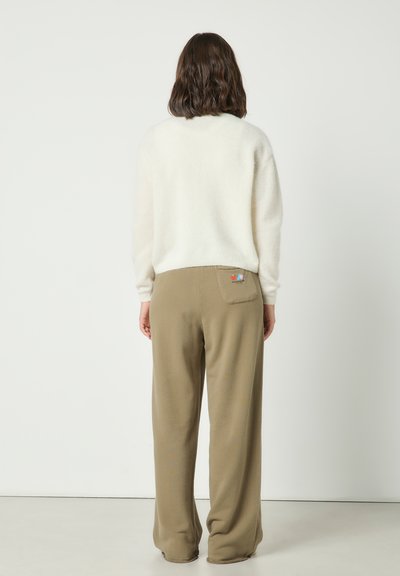Femme aux cheveux bruns mi-longs portant un pull blanc et un pantalon ample kaki avec un petit écusson coloré brodé sur la poche arrière.