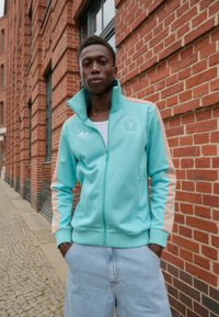 Mintgrøn zip-up jakke med orange striber, ribbede manchetter og et emblem på venstre bryst. Kombinationen med lyseblå denimbukser.