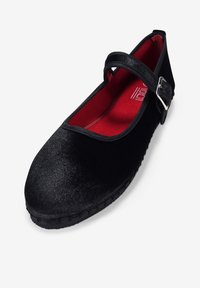 Zapato plano negro con punta redonda, parte superior de tela texturizada y una sola correa ajustable con hebilla plateada. Forro interior rojo.