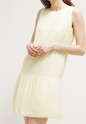 Ärmelloses blassgelbes Kleid mit ausgestelltem Saum, getragen von einer Frau, gezeigt von Hals bis zu den Knien mit locker verschränkten Händen.