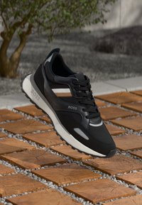 Czarne buty sportowe z siateczkową cholewką, kontrastującymi paskami w beżowym i białym kolorze oraz białą podeszwą środkową, stojące na teksturowanych brązowych kostkach brukowych.
