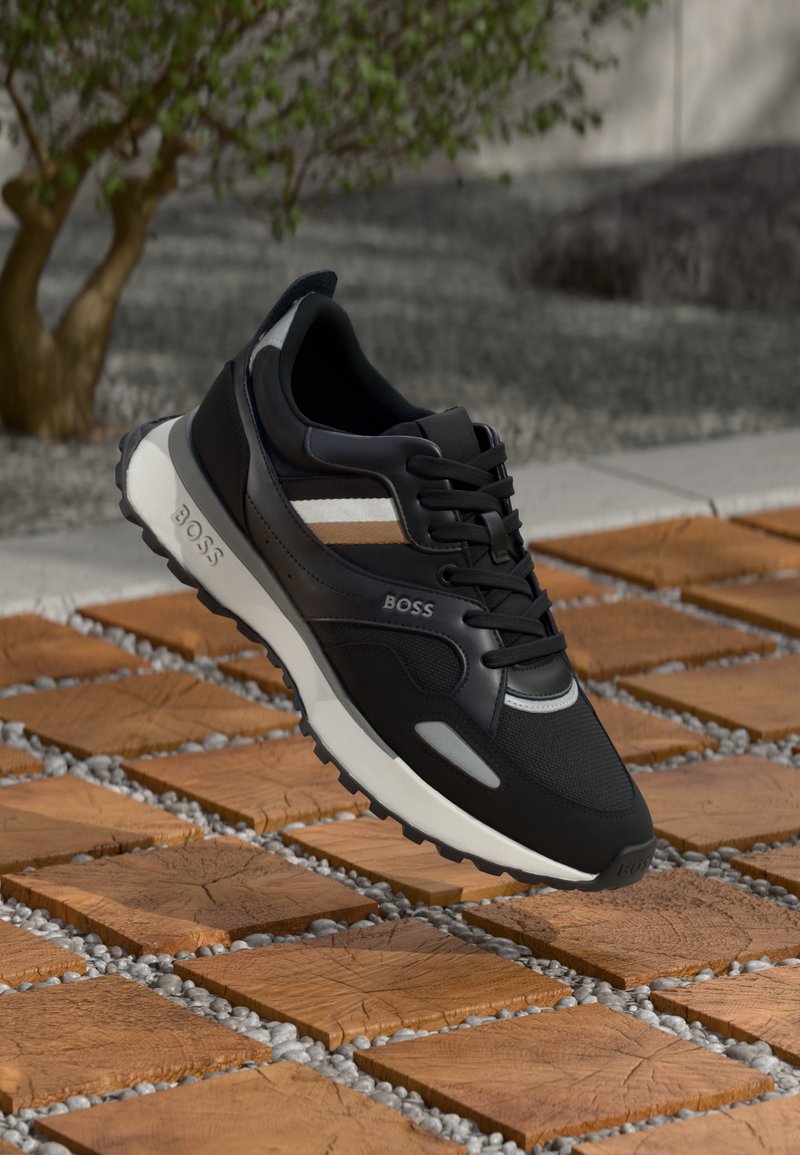 Czarne buty sportowe z siateczkową cholewką, kontrastującymi paskami w beżowym i białym kolorze oraz białą podeszwą środkową, stojące na teksturowanych brązowych kostkach brukowych.