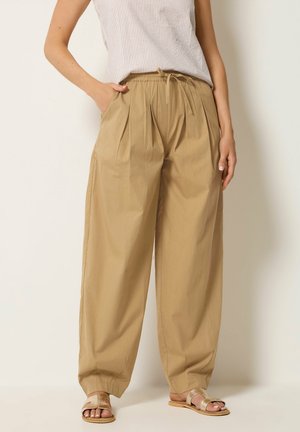 Personne portant un pantalon beige ample à plis, un haut sans manches à rayures, des sandales dorées et les mains dans les poches, debout devant un fond uni.