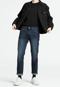 Veste en denim noire sur un t-shirt noir, avec un jeans bleu foncé retroussé au niveau des chevilles. Des baskets blanches complètent la tenue. Look simple et décontracté.