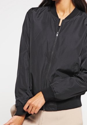 Frau trägt eine schwarze Bomberjacke mit Reißverschluss, geripptem Kragen, Bündchen und Saum und richtet den Jackenärmel.