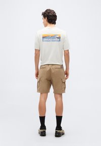 Άνδρας που στέκεται με την πλάτη στραμμένη, φοράει λευκό T-shirt Patagonia, μπεζ cargo σορτς, μαύρες κάλτσες και μαύρα αθλητικά παπούτσια Salomon σε λευκό φόντο.