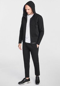 Czarna kurtka zapinana na zamek błyskawiczny z kapturem, z dwoma przednimi kieszeniami i purpurową podszewką, w zestawieniu z czarnymi spodniami zwężającymi się ku dołowi i czarnymi sneakersami. Pod spód biała koszulka.