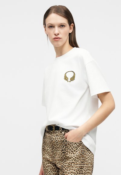 Γυναίκα που φοράει ένα λευκό t-shirt με δύο τίγρεις που σχηματίζουν έναν κύκλο στο στήθος, συνδυασμένο με παντελόνι με λεοπάρ μοτίβο και μαύρη ζώνη.